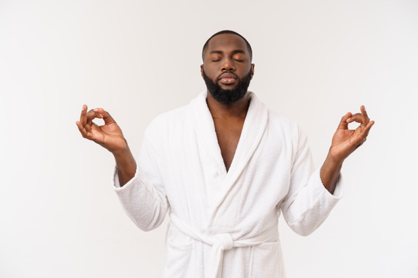 Gedachte Afro-Amerikaanse man in badjas kalmerend, diep ademend, mediterend met gesloten ogen, mudra gebaar, jonge man die yoga beoefent, oefening doende, geïsoleerd staand op studio achtergrond - Foto, afbeelding