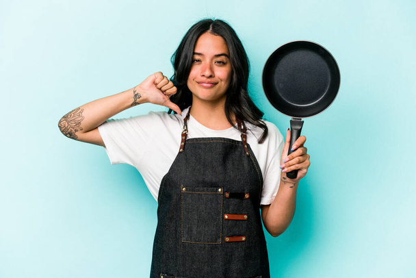 Young hispanic cooker woman holding frying pan isolated on blue background feels proud and self confident, example to follow. - 写真・画像