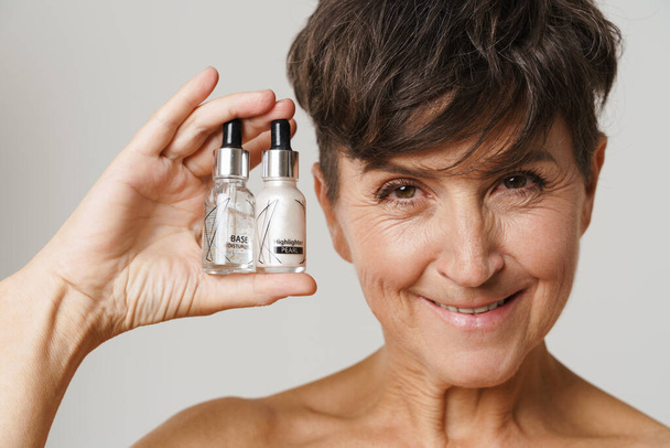 Mature half-naked woman smiling while showing cosmetic products isolated over white background - Φωτογραφία, εικόνα