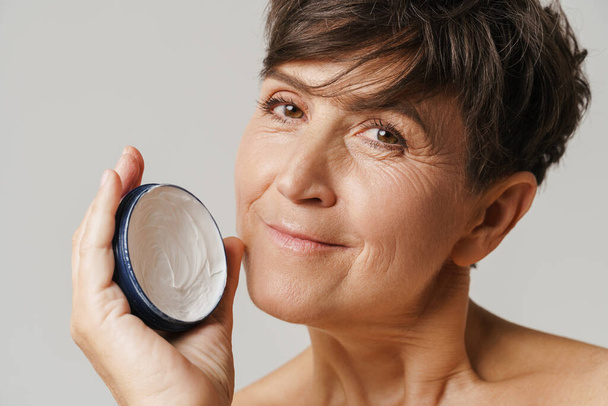 Mature half-naked woman smiling while showing face cream isolated over white background - Photo, image