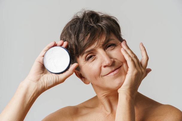 Mature half-naked woman smiling while applying face cream isolated over white background - Fotó, kép