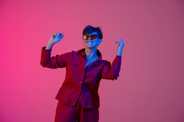 Mature european woman wearing sunglasses dancing on camera isolated over pink background - Фото, изображение