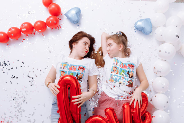 Του Αγίου Βαλεντίνου. Η νεαρή μητέρα και η έφηβη κόρη της διασκεδάζουν και κρατούν κόκκινη αγάπη και μπαλόνι σε σχήμα καρδιάς ξαπλωμένο σε εορταστικό φόντο με κομφετί. Ημέρα μητέρων. Πάρτι γενεθλίων - Φωτογραφία, εικόνα