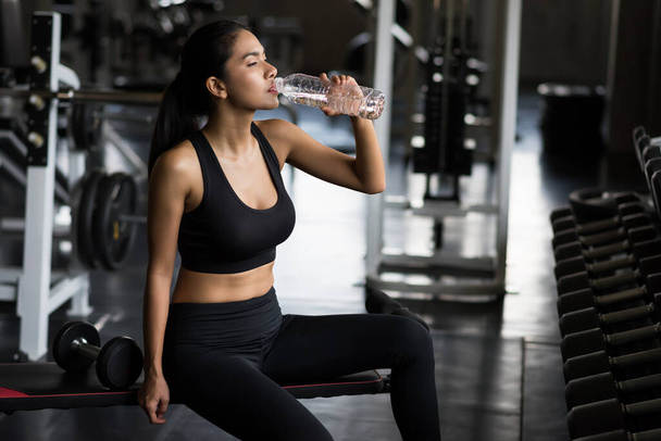 Fit Asiatinnen trinken Wasser und ruhen sich nach der Hantelübung im Fitnessstudio aus. Asiatisches Mädchen macht eine Pause und entspannt sich auf dem Sportbankstuhl. Bodybuilding und gesunder Lebensstil. - Foto, Bild