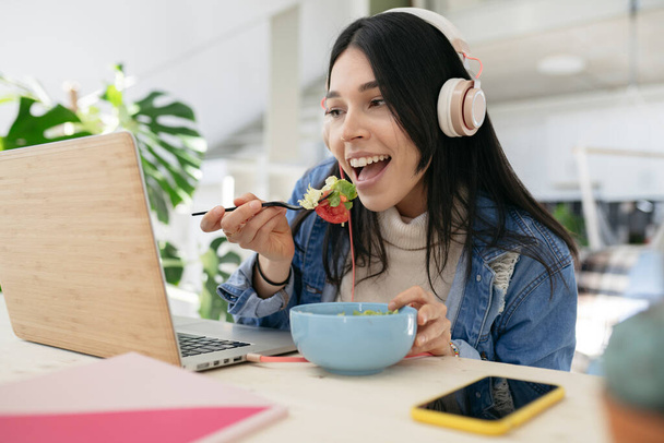 bella donna a casa con le cuffie utilizzando il computer portatile e mangiare insalata fresca - Foto, immagini
