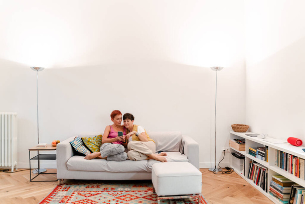 Mature lesbian couple using mobile phone while resting on sofa together at home - Valokuva, kuva