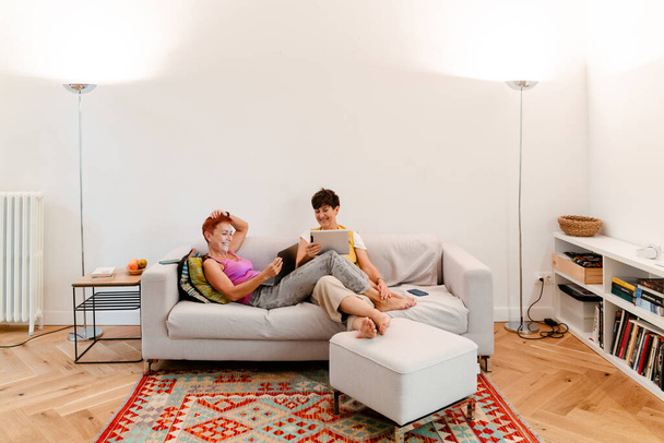 Mature lesbian couple using gadgets while resting on sofa together at home - 写真・画像