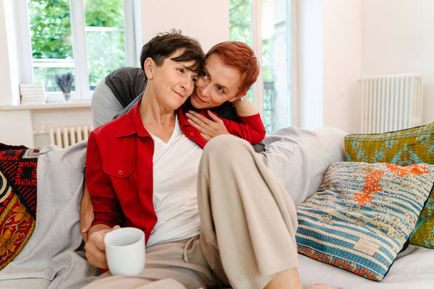 Mature lesbian couple hugging and drinking coffee on sofa at home - Фото, зображення