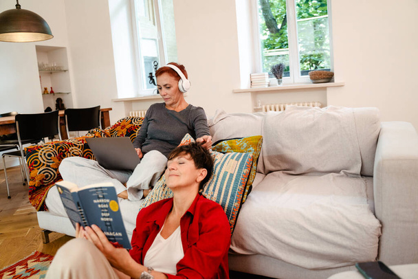 Mature lesbian couple using laptop and reading book at home - 写真・画像