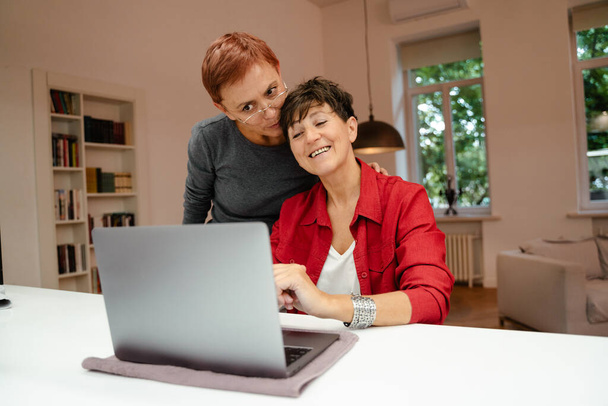 Mature lesbian couple smiling and hugging while using laptop at home - Fotó, kép