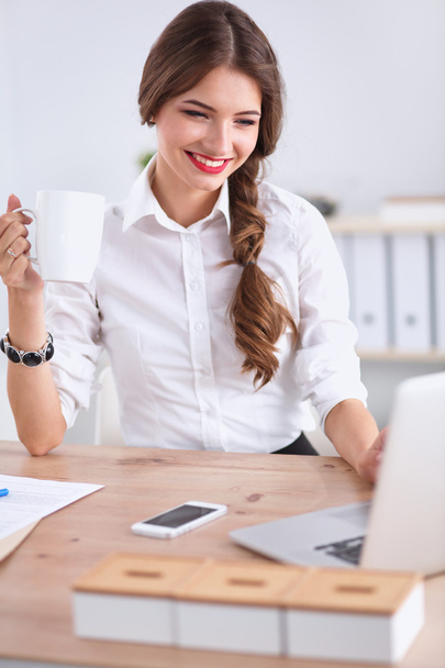 Young businesswoman sitting on the desk with cup in office - Фото, изображение