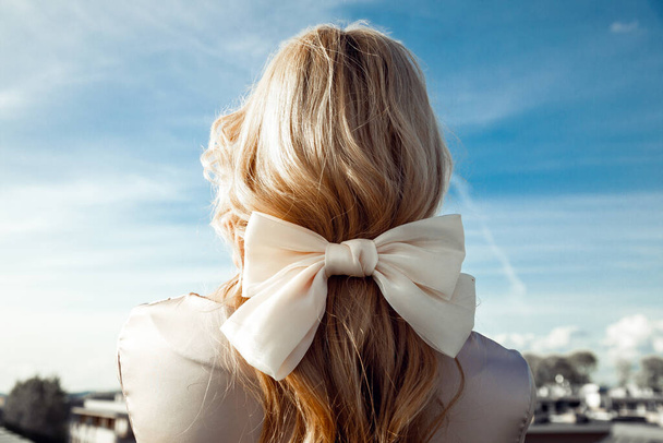 Vue de dos de la femme avec de longs cheveux clairs décorés avec un arc beige en ruban debout sur fond bleu ciel. Gros plan - Photo, image