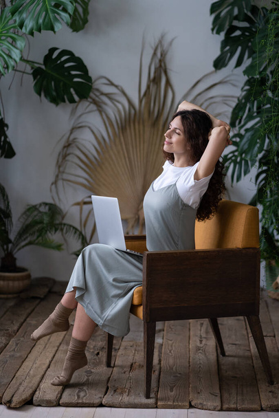 Beautiful woman with hands behind head and computer on knees resting, daydreaming in surround plants - Photo, image