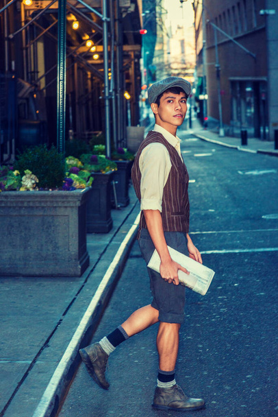 Chłopiec z miasta. Noszenie czapki newsboy, koszula, kamizelka wzorzyste, szare spodnie, buty buty, trzyma gazetę, Asian American student chodzenia, przejście wąskiej ulicy w Nowym Jorku Cit - Zdjęcie, obraz