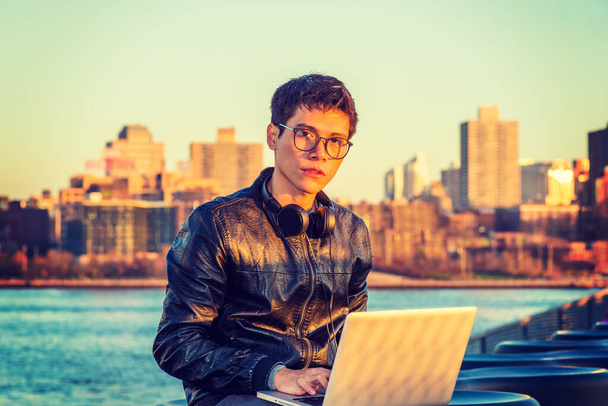 Asiatisch-amerikanischer College-Student auf Reisen, studiert in New York, trägt schwarze Lederjacke, Brille, Kopfhörer im Nacken, sitzt bei Sonnenuntergang am Fluss, arbeitet am Laptop.  - Foto, Bild