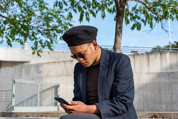 A fiatal latin meleg férfi divatos kalapot és napszemüveget visel, SMS-ezik az okostelefonján, miközben a tengerparton üldögél a szabadban. LGTB.  - Fotó, kép