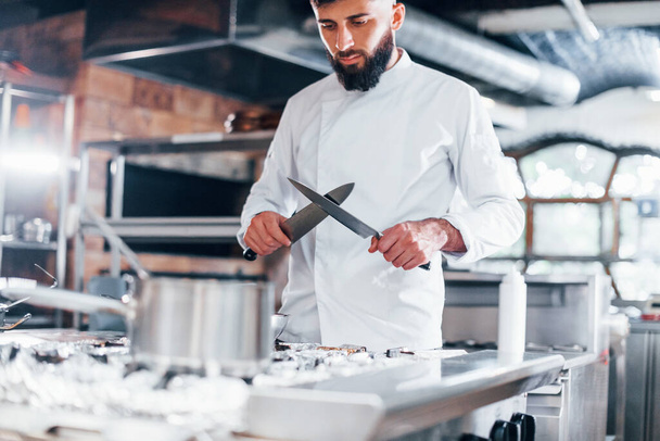 Chef in white uniform standing at kitchen. Holding knives in hands. - Foto, Bild