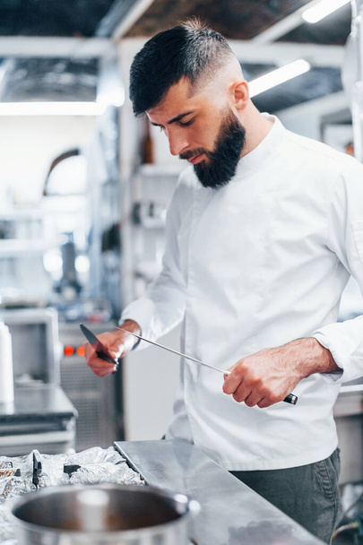 Chef in white uniform standing at kitchen. Holding knives in hands. - 写真・画像