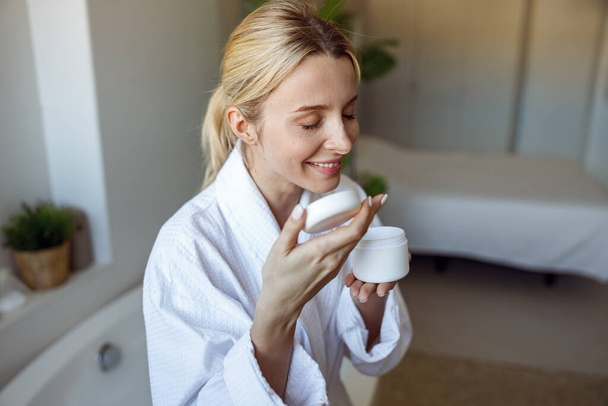 Attractive young Caucasian female smelling facial cream or body lotion in bathroom. - Φωτογραφία, εικόνα