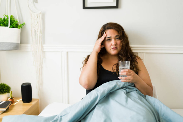 Πορτρέτο μιας θλιμμένης παχύσαρκης γυναίκας με ημικρανία που υποφέρει από πονοκέφαλο και πίνει ένα ποτήρι νερό στο κρεβάτι  - Φωτογραφία, εικόνα