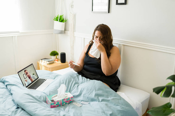 Mujer joven enferma con obesidad llorando mientras escucha el diagnóstico médico de un médico virtual durante una videollamada en la cama  - Foto, imagen