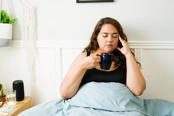 Mujer triste de 20 años con obesidad que sufre de dolor de cabeza o migraña mientras bebe té caliente en la cama - Foto, Imagen