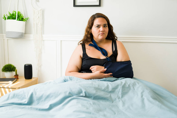 Portret van een trieste depressieve dikke vrouw met een gebroken arm die in haar bed rust in pyjama 's en een gips gebruikt vanwege een blessure - Foto, afbeelding
