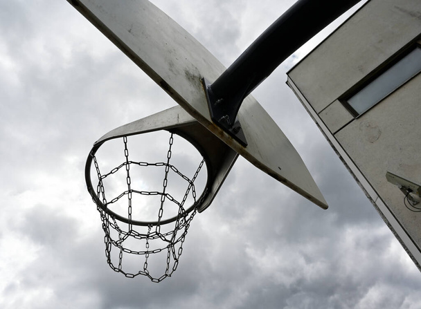 um aro de basquete anti-vândalo com correntes de ferro contra um céu nublado sombrio, ao lado de um edifício de concreto com uma câmera de vigilância na parede  - Foto, Imagem