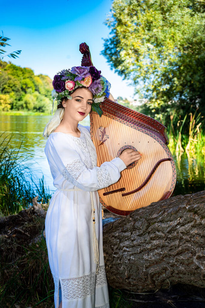 Young Ukrainian woman in national dress plays a bandura by the river on a summer sunny day. - Φωτογραφία, εικόνα