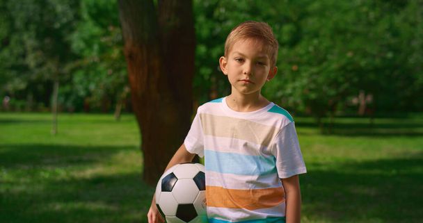 Chłopiec z piłką nożną pozowanie kamery na przyrodę. Poważny mały sportowiec zbliżenie. - Zdjęcie, obraz