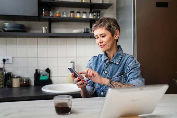 One woman mature caucasian female using mobile phone in the kitchen in the morning daily routine real people leisure and relaxation concept copy space making a call talking happy smile - Foto, Bild