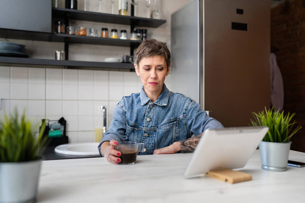 jedna kobieta dojrzała kaukaska kobieta posiadające filiżankę kawy w kuchni podczas korzystania z tabletu cyfrowego codziennie rano rutynowe sprawdzanie poczty lub codziennych zadań podczas przygotowywania się do pracy prawdziwi ludzie skopiować przestrzeń - Zdjęcie, obraz
