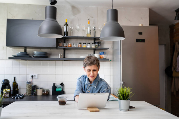 one woman mature caucasian female having a cup of coffee in the kitchen while using digital tablet daily morning routine checking mail or daily tasks while preparing for work real people copy space - Photo, image