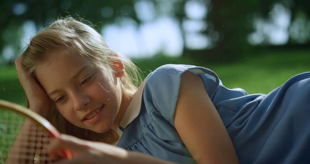 Menina sorrindo deitado no gramado verde sozinho. Loira criança toque raquete net closeup - Foto, Imagem