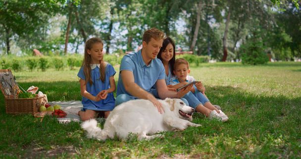 White dog lie down near family holding tablet on picnic. Father caress labrador. - Photo, Image