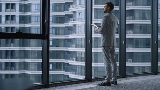 Pensando hombre ejecutivo de pie utilizando tableta de trabajo en línea en la oficina de la ventana. - Foto, Imagen
