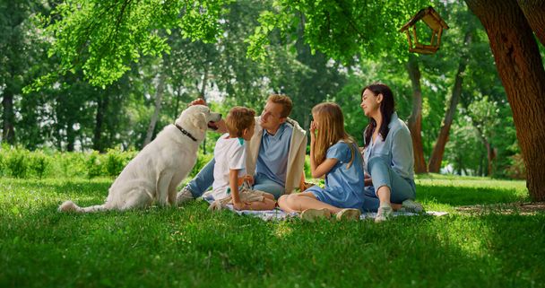 Νεαρή οικογένεια χαϊδεύει λαμπραντόρ στο πικνίκ. Ευτυχισμένοι άνθρωποι ξεκουράζονται με το σκυλί έξω. - Φωτογραφία, εικόνα