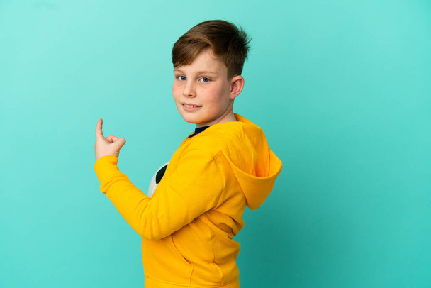 Redhead kid playing football isolated on blue background pointing back - Valokuva, kuva