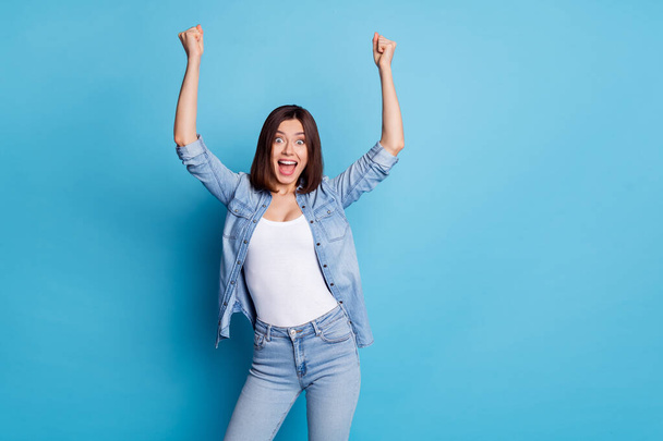 Photo of shiny lucky woman wear jeans shirt rising fists screaming empty space isolated blue color background - Photo, Image