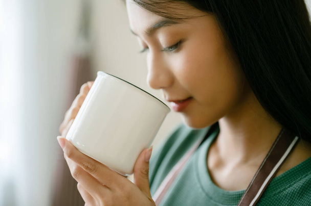 Jeune femme asiatique debout à côté de la fenêtre et tenant une tasse dans la salle de cuisine à la maison, elle boit du lait après le réveil le matin - Photo, image