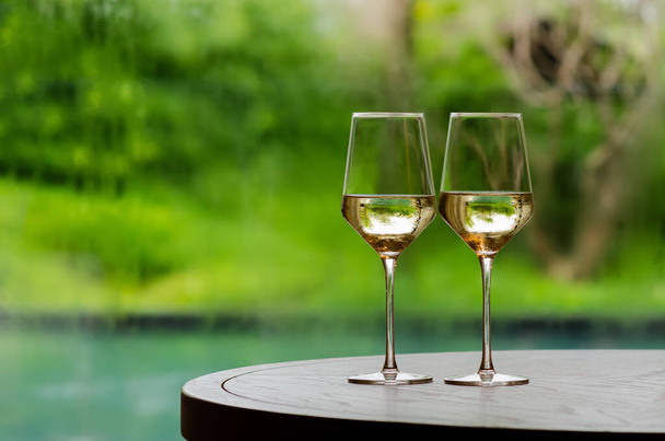 木製のテーブルの上に白ワインの2つのガラスは、ぼやけたスイミングプールと庭の背景を持つカップルのために祝うために. - 写真・画像