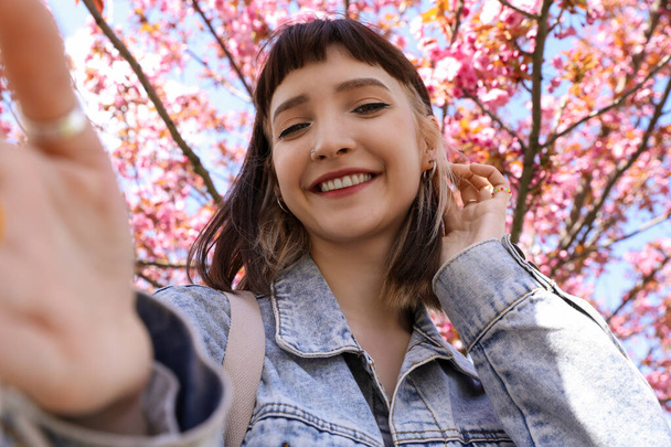 Beautiful young woman taking selfie in park with blossoming sakura trees - Foto, afbeelding