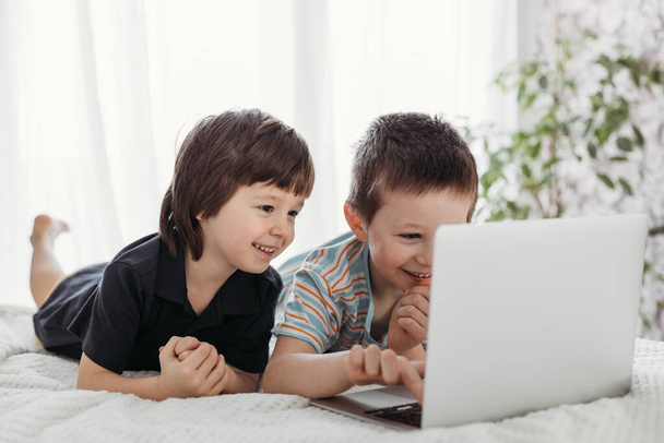 Enseñanza a distancia en casa. Los niños aprenden usando tabletas digitales y computadoras portátiles. Los chicos están viendo algo en la computadora mientras están acostados en la cama.. - Foto, Imagen