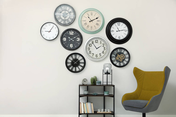 Armchair, console table and many different clocks hanging on white wall in room - Foto, imagen