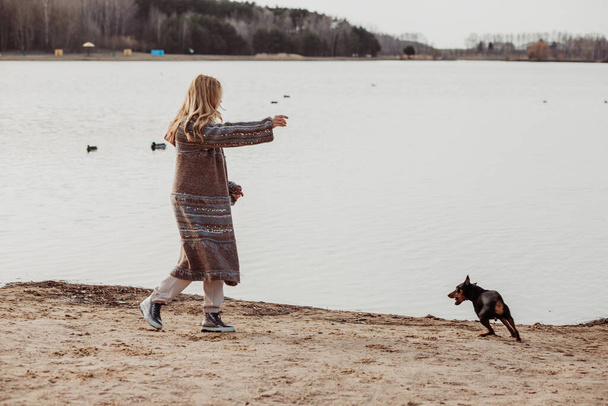 Happy enjoying blonde woman in warm clothes playing and having fun with cute puppy near river bank on cool sunny day, ducks swimming in water. Outdoors weekend activity, dogginess, water background - Photo, Image