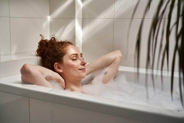 Fiatal vörös hajú nő vesz egy fürdőt csukott szemmel  - Fotó, kép