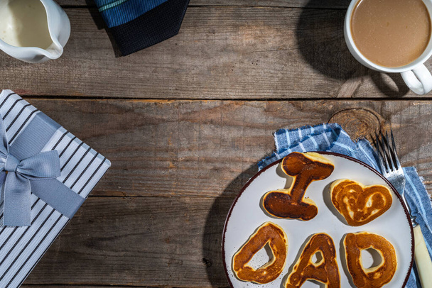 Father day pancakes. Homemade kids cooked father day pancakes with lettering I love dad, on plate, with coffee latte cup, gift box and necktie, wooden background top view copy space - Foto, afbeelding