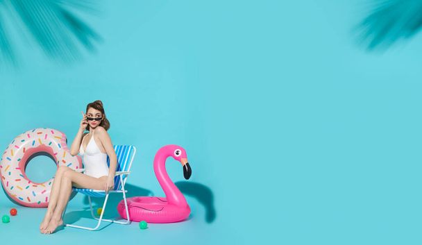 Vrolijke vrouw chillen in lounge stoel in de buurt van flamingo rubberen ring op blauwe kopieerruimte achtergrond. vrouw ontspannen op het strand in de zomer vakantie. - Foto, afbeelding