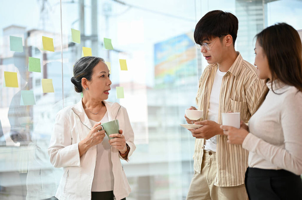 Onnistunut aasialainen vanhempi naispuolinen pomo puhuu ja jakaa luovan idean kahden nuoren työntekijän kanssa toimistossa kahvitauolla.. - Valokuva, kuva