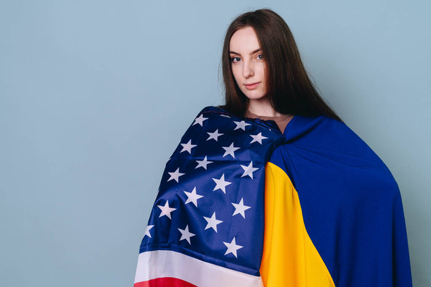 Retrato de close-up de uma menina com cabelo castanho em um fundo azul com a bandeira americana e ucraniana. O conceito de apoio dos EUA à Ucrânia. - Foto, Imagem